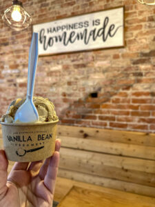 Butter pecan ice cream in a cardboard cup labelled Vanilla Bean Creamery with a "Happiness is Homemade" sign behind